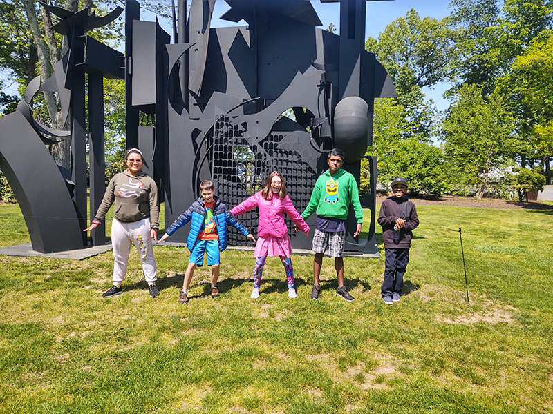 A large black sculpture is in the background and five middle school students stand in front of it with their arms outstretched.