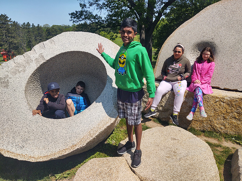 Five middle school students sit or stand on rocks. Some are hollowed out and kids are sitting in them. There is a blue sky in the background.