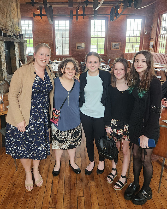 A woman with long blonde hair on the left, wearing a blue print dress and beige jacket, with four middle school girls. all are smiling and dressed nicely.