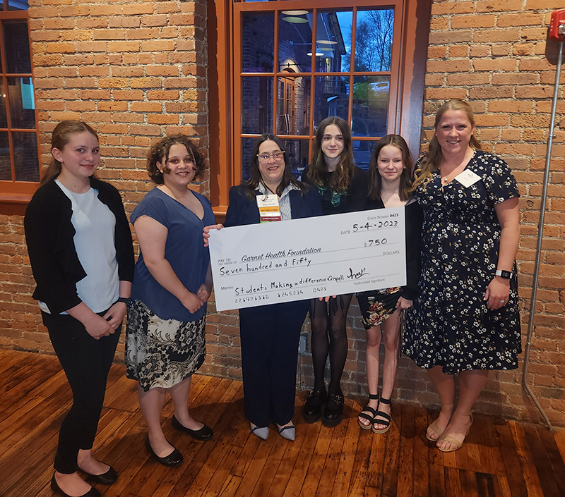 A woman with glasses and long dark hair in the center holds a large check for $750. There are two young women on the left and two young women on the right. Far right is a woman with long blonde hair wearing a blue printed dress. All are smiling.