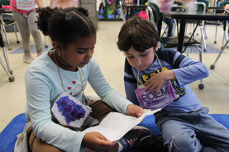 Two first-grade students sit on the floor together. The boy on the right is pointing to the paper the girl on the left is reading from.