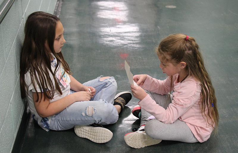 Two first grade girls sit cross-legged on the floor. The girl on the left reads her poem from a piece of paper to the girl on the left.