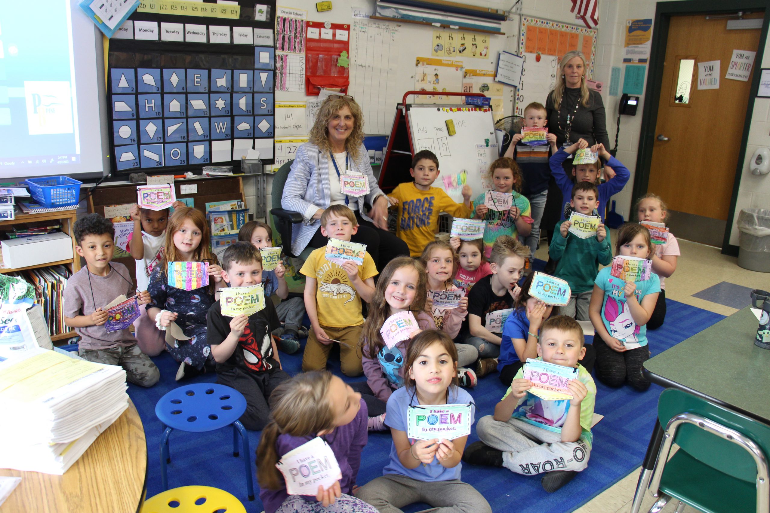 A class of first graders sitting on the floor hold up their pockets, which they made from paper. In the back are two women also with their pocket.