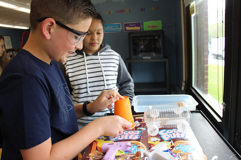Two fifth grade students work on creating a vehicle. The kidon the left is wearing safety goggles and cutting an orange cup. There is another student standing watching.