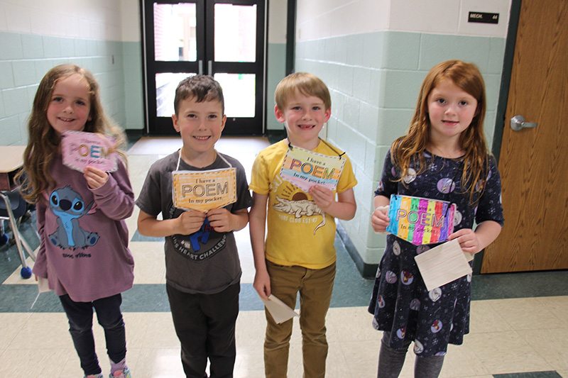 Four first-grade students smile and hold up their paper pocket, which is on a string around their necks. The pockets have their poems in them.