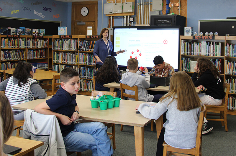 A woman stands in front of a class pointing to a white board while students sit at a table  watching and listening.