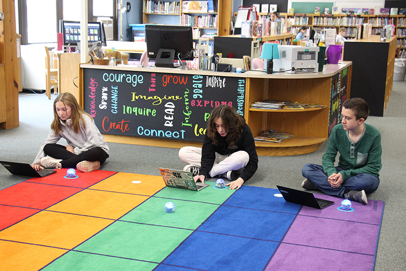Three fifth-grade students sit at the edge of a multi-colored rug. They are sitting with their Chrome books programming the sphere in front of them.