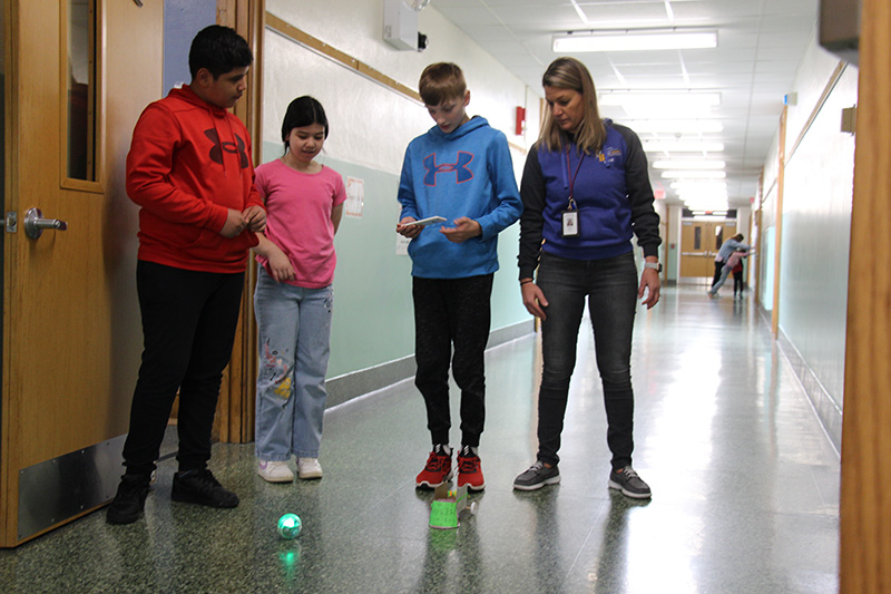 Three fifth-grade students stand in a hallway with a teacher. One student has a phone in his hand and is controlling the movements of the lit sphere on the floor.