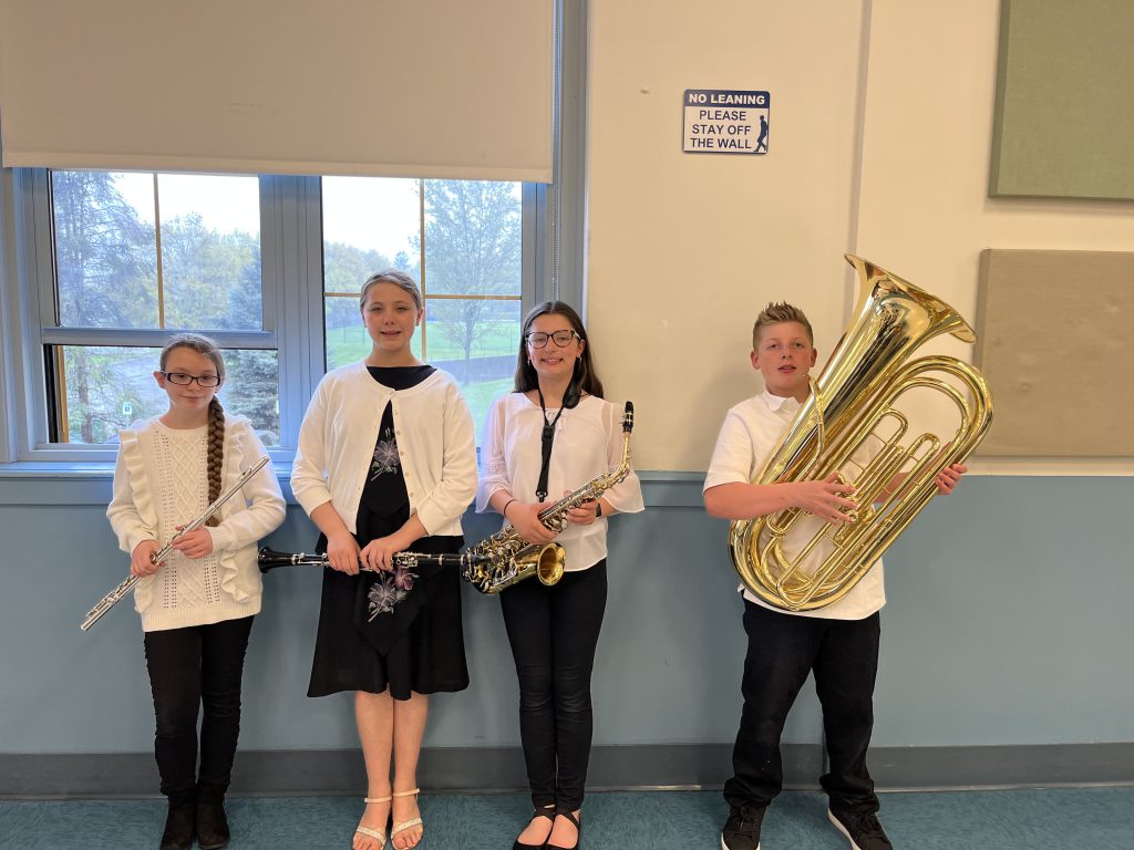 Four middle school age students stand together holding instruments. Three girls and one boy on the right. He is holding a large tuba while the others have flutes and clarinets.