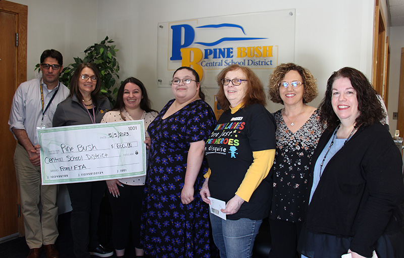 Seven adults stand in front of a blue and gold Pine Bush Central School District sign. The three people on the left have a large ceremonial check made out to the district for $600.