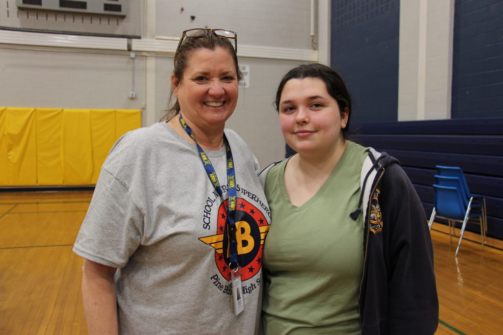 A woman on the left is wearing a gray tshirt with blood drive information on the front. She is smiling. Her long hair is pulled back in a ponytail and she has glasses on top of her head.  On the right is a high school student with dark hair and wearing a green shirt. She is smiling.