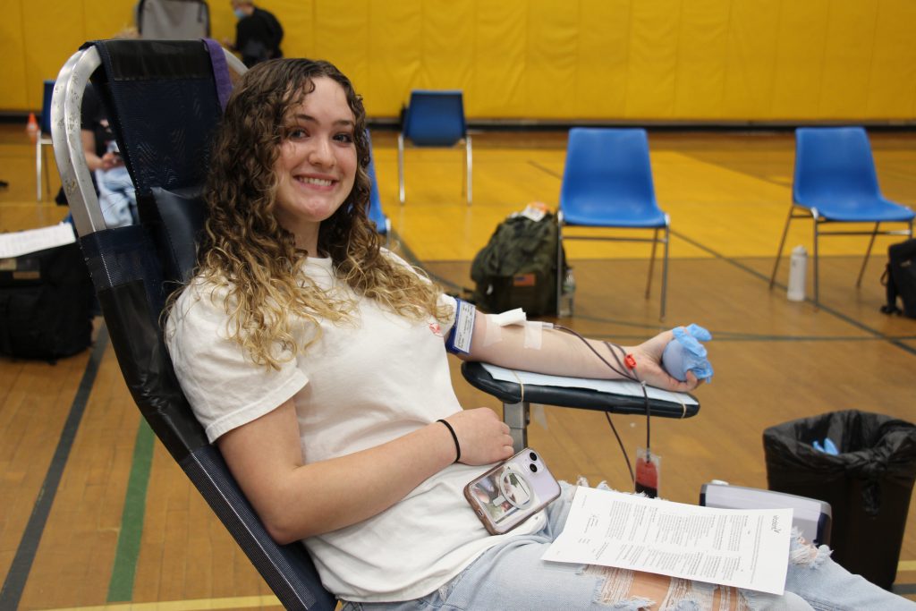 A young woman sits reclined on a chair as she donates blood. She is smiling.