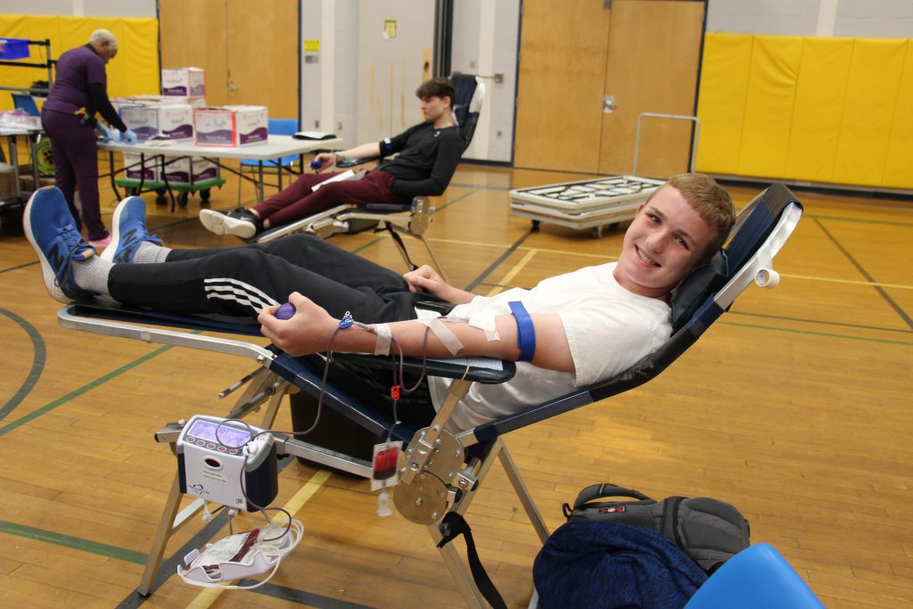 A young man sits reclined as he donates blood in a gymnasium. He is smiling.