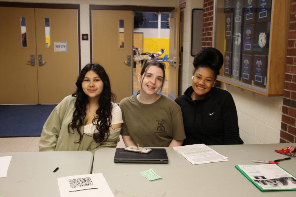 three young women sit at a table where they are checking in people for a blood drive.