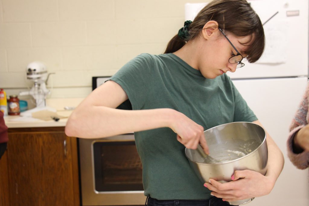 A high school girl with her dark hair pulled back in a ponytail  mixes batter in a stainless steel bowl.