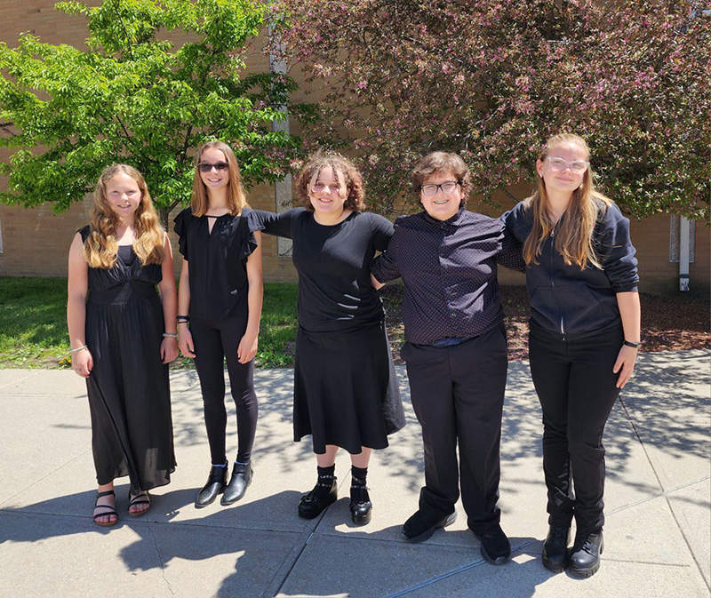 Five middle school students, four girls and one boy all dressed in black, stand arm-in-arm outdoors on a beautiful sunny day.