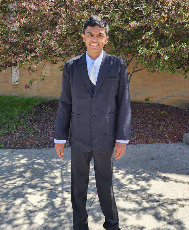 A middle school boy dressed in a dark suit with a blue shirt and tie, stands outside on a beautiful day.