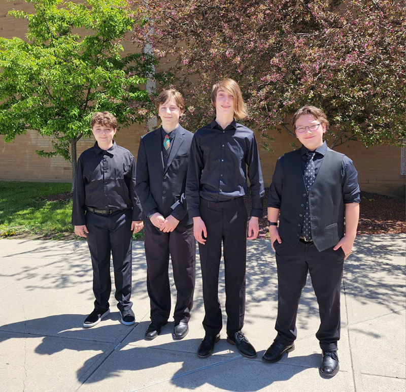 Four boys dressed in black shirts and pants, stand outside on a beautiful day.