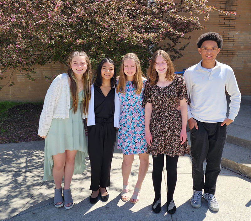 Five middle school students - four girls and one boy, stand closely and smile. They are outside on a beautiful day.