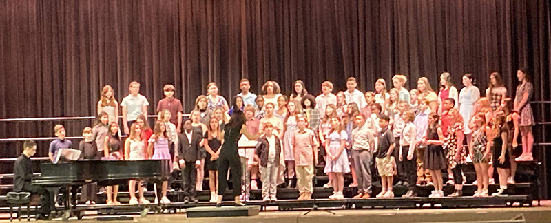 A large group of middle school students are on stage singing. There is a student playing piano off to the left and a woman directing.