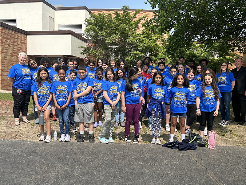 A group of about 30 elementary age students, all wearing blue shirts, and three adults on the sides stand in front of a building and tree.