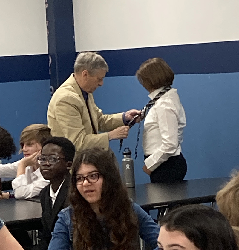 A man on the left, wearing a shirt and tie, ties the tie of a middle school student, who is wearing a white shirt. Other students are around them.