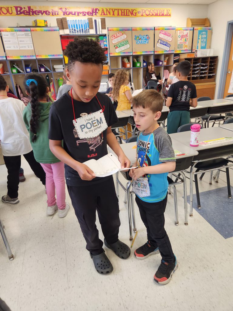 Two second-grade boys stand together with the boy on the left reading a poem from a piece of paper to the boy on the right.