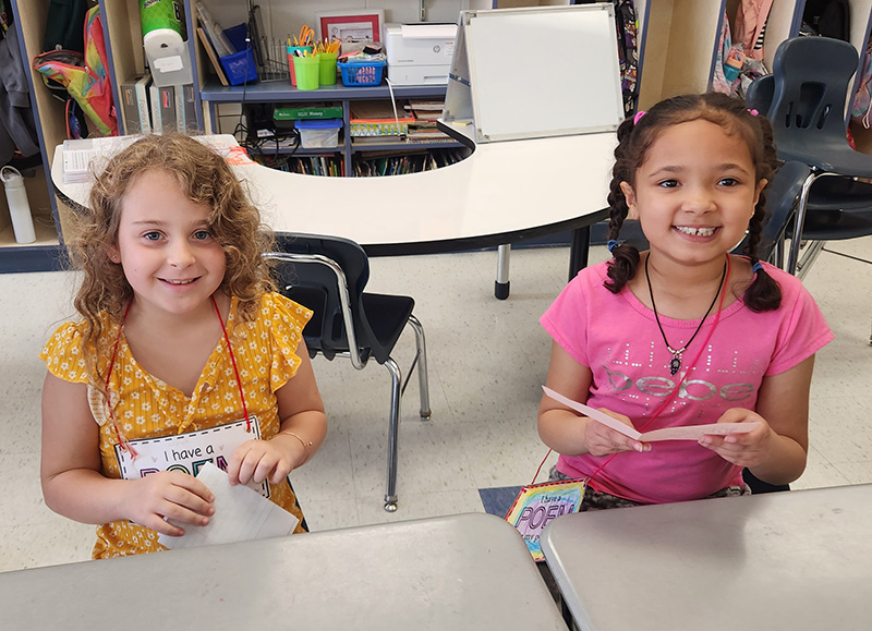 Two girls sit at desks smiling, holding their poems in their pockets.