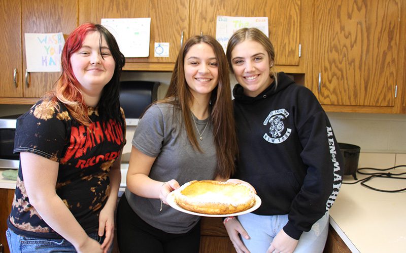 Three high school girls smile as they hold a large fluffy pancake on a plate in front of them.
