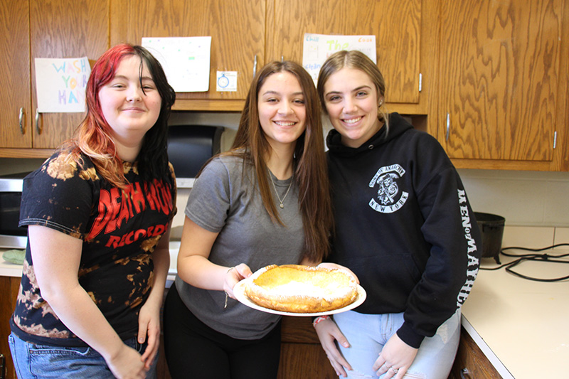 Three high school girls smile as they hold a large fluffy pancake on a plate in front of them.