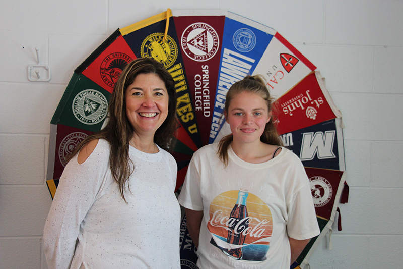 A woman wearing a white shirt with long dark hair smiles as she stands with a young woman with her hair in a ponytail, wearing a white tshirt. Behind them are colorful college pennants in the shape of a circle.