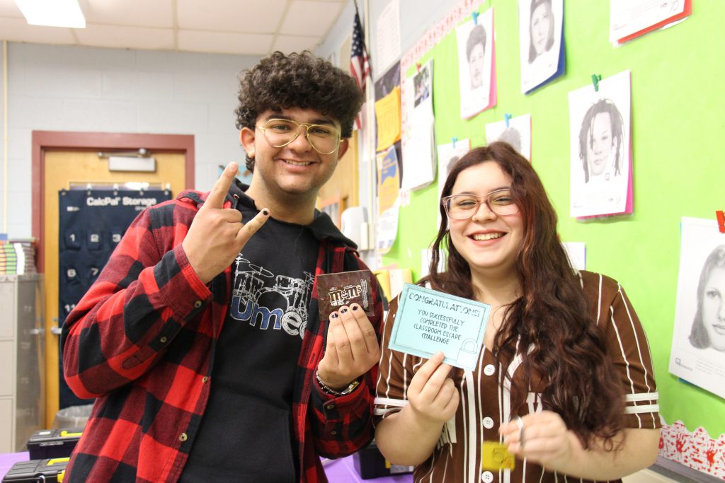 two high school students, a boy on the left wearing a red and black plaid shirt,  and a girl on the right wearing a brown baseball jersey, smile. The boy is holding up a candy bar and the girl is holding up a card saying you won.