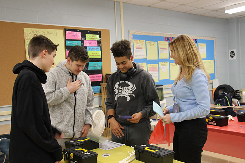 Three high school boys, all wearing black or gray hoodies, work on an escape problem with a woman with long blonde hair.