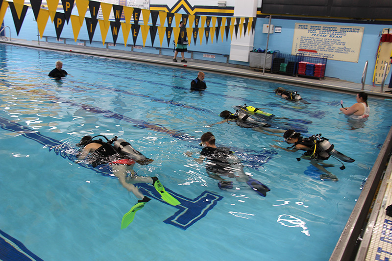 A pool with five high school students in scuba gear snorkeling on top of the water. There are people standing in the water watching.