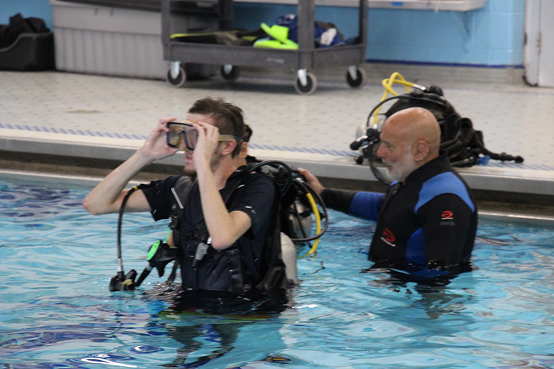 A high school age young man puts goggles on in a pool while a man behind him helps adjust an air tank on his back.