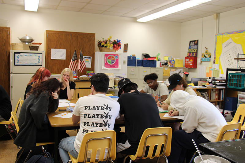 A group of high school kids sit around a table doing paperwork.