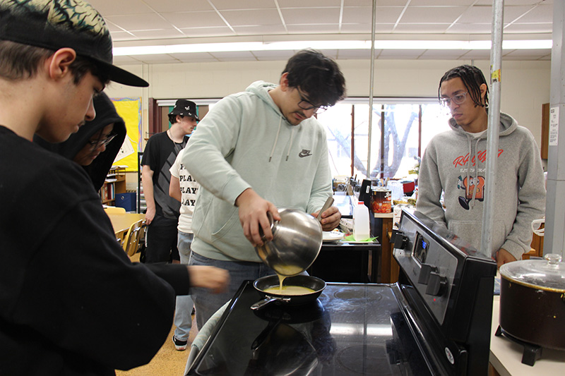 A group of four high school boys pour their batter into a pan on a stove.