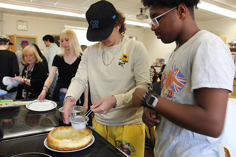 Two high school boys sprinkle powdered sugar on a large fluffy pancake.