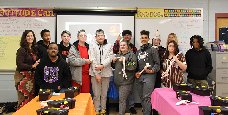 A group of 11 high school students and two women stand in front of a classroom smiling. Some of the kids are holding items from their forensics class like a skeleton.