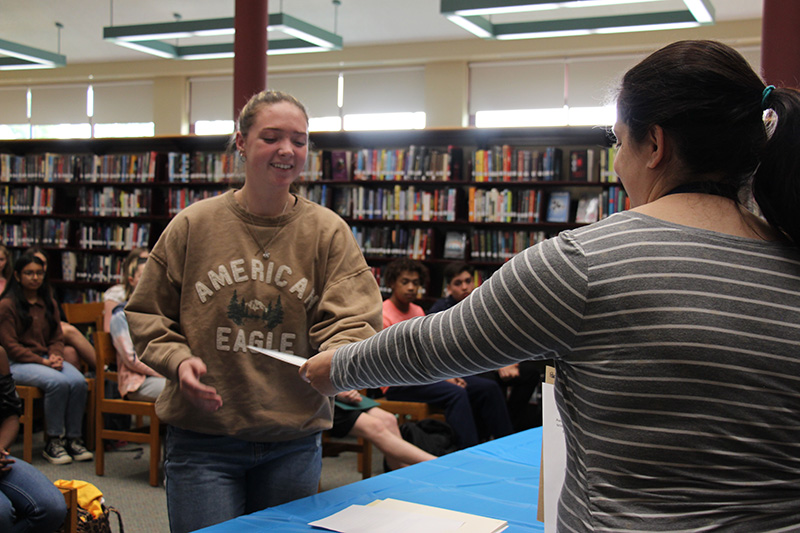 A high school girl in tan sweatshirt and her long hair pulled back smiles as she accepts an award from a woman in a black and white striped shirt.