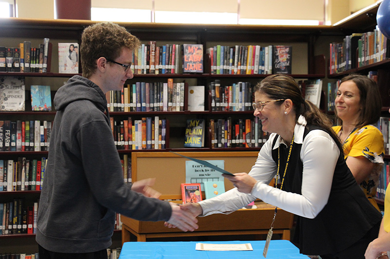 A high school boy with glasses wearing a dark colored hoodie shakes hands with a woman and accepts an award form her.