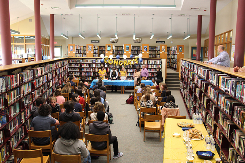 A view of a room from above. There are high school students sitting on wooden chairs. In the front of the room is a long table with five adults standing at it. It is covered in a blue table cloth.