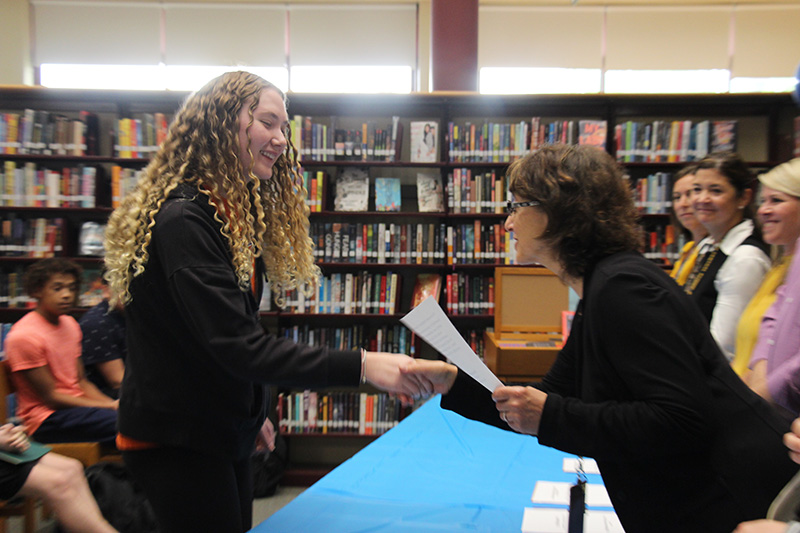 A high school girl with long blonde hair shakes hands with a woman and accepts an award from her.