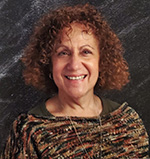 A woman with curly brown hair smiles. She is wearing a brown and black sweater.