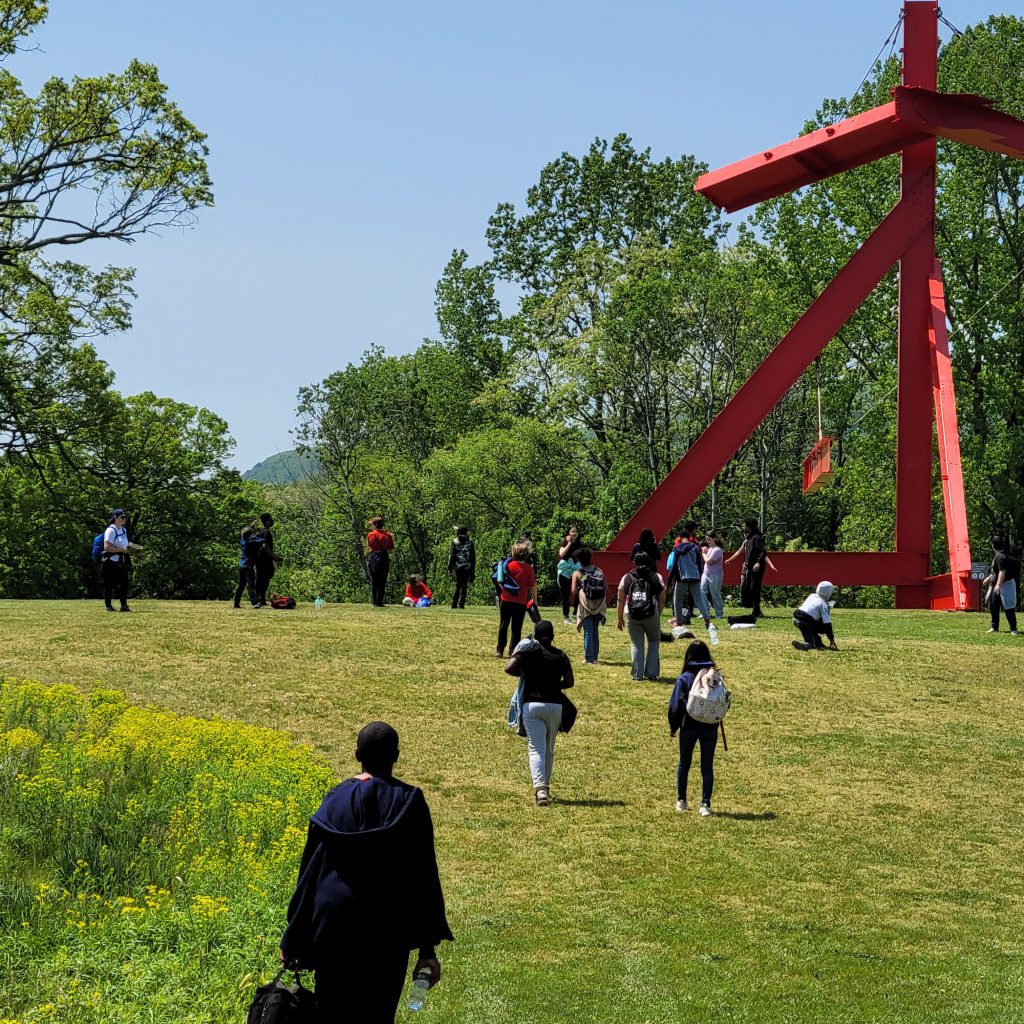 A large orange sculpture in the grass. There are many middle school students walking toward it.