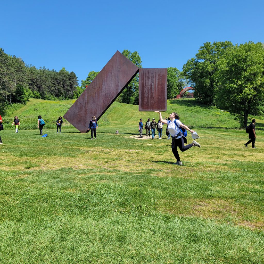 A large two-piece metal sculpture in a field of grass. There is a middle school girl in front positioned like she is holding it up.