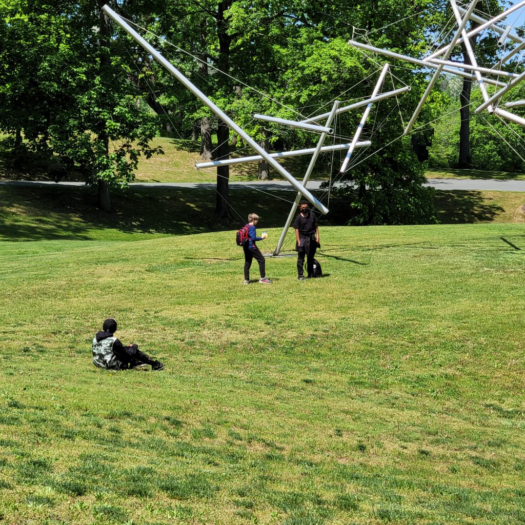A large metal sculpture n a grassy field with students around it.