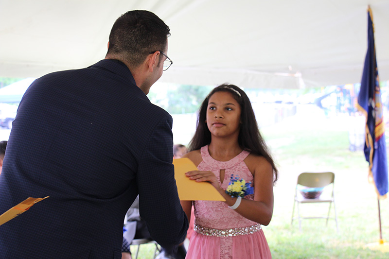 A fifth-grade girl in a pink dress with long dark hair accepts an envelope from a man in a suit and shakes his hand.