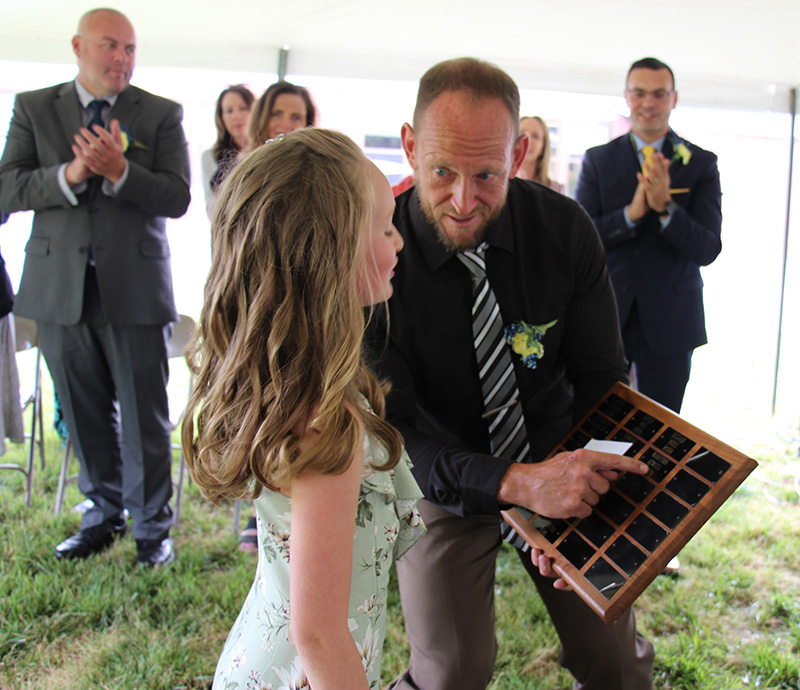 A fifth-grade girl with long blonde hair looks at a plaque which is held by a man in a dark shirt and striped tie.