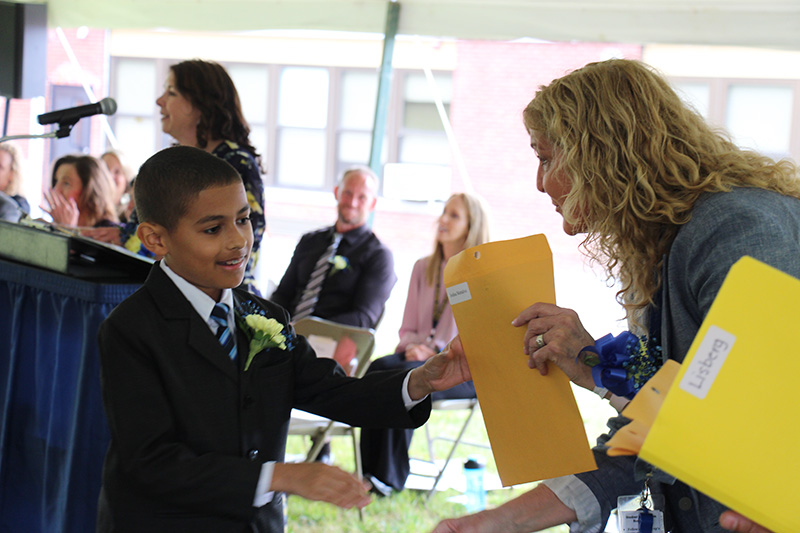 A young man in a suit and tie shakes hands with a woman with long blonde hair. She is handing him an envelope.